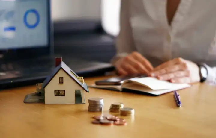 A model house beside stacked coins and a notebook, charts on the screen behind